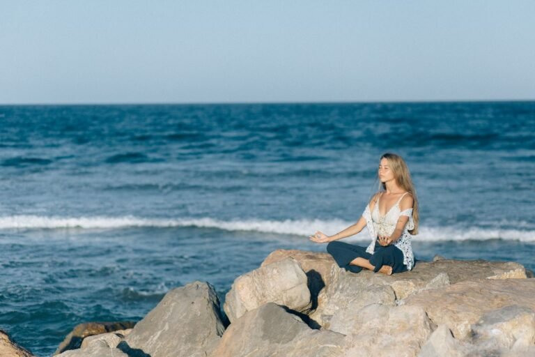 A woman practices meditation outdoors on rocks by the sea, embodying tranquility and relaxation.